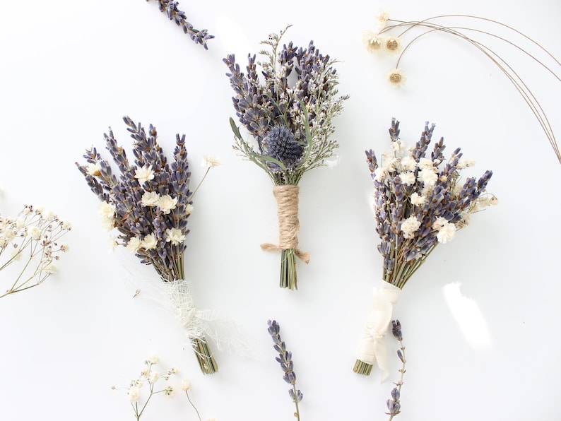 May include: Three small bouquets of dried lavender and white flowers tied with twine and ribbon. The bouquets are arranged on a white surface.