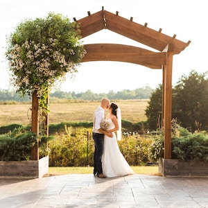 May include: A couple embraces and kisses under a wooden arbor, framed by a field and trees. The bride wears a white wedding dress and veil, holding a bouquet. The groom wears a vest, white shirt, and jeans. A flowering vine decorates a wooden post.