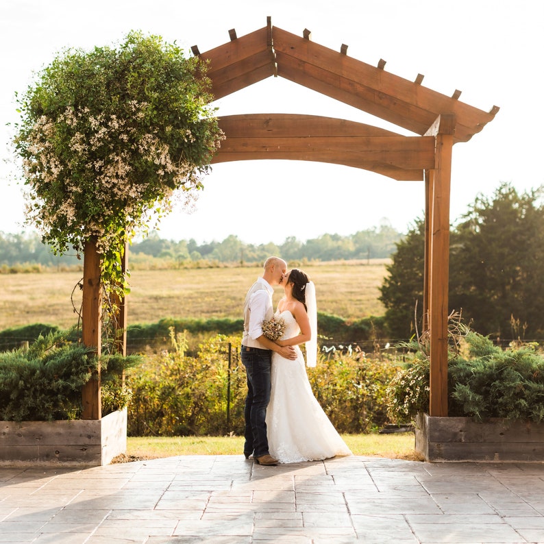May include: A couple embraces and kisses under a wooden arbor, framed by a field and trees. The bride wears a white wedding dress and veil, holding a bouquet. The groom wears a vest, white shirt, and jeans. A flowering vine decorates a wooden post.