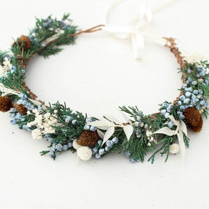 May include: A handcrafted floral crown featuring evergreen sprigs, blue berries, white accents, and small pinecones. The crown is tied with a white ribbon, perfect for weddings or special occasions.