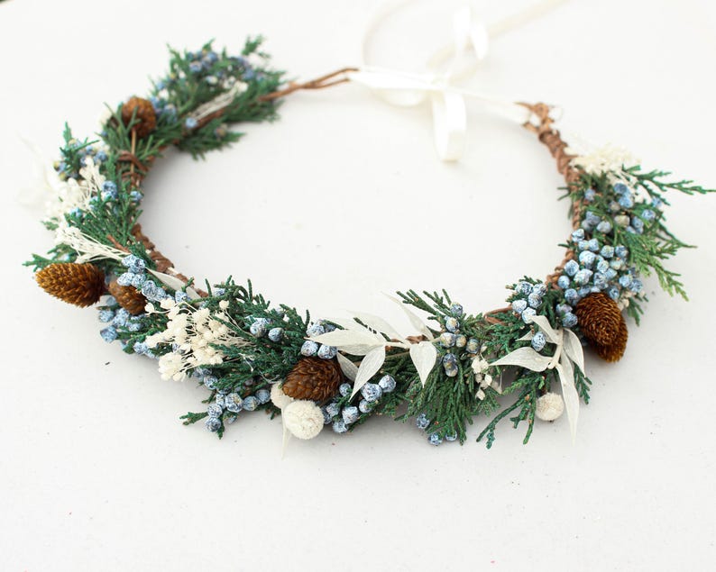 May include: A handcrafted floral crown featuring evergreen sprigs, blue berries, white accents, and small pinecones. The crown is tied with a white ribbon, perfect for weddings or special occasions.