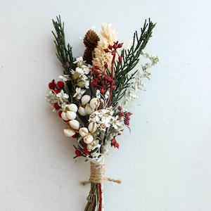 May include: A small bouquet of dried flowers and greenery. The arrangement includes white, red, and beige flowers, with green sprigs and a brown pinecone. The stems are bound with twine, and the bouquet is set against a white background.