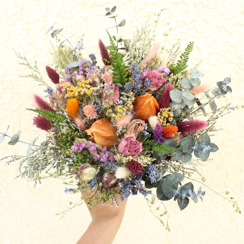 May include: A vibrant bouquet of dried flowers in shades of purple, pink, orange, and green. The arrangement includes roses, eucalyptus, and various other dried plants, creating a textured and colorful display. The bouquet is held in a hand.
