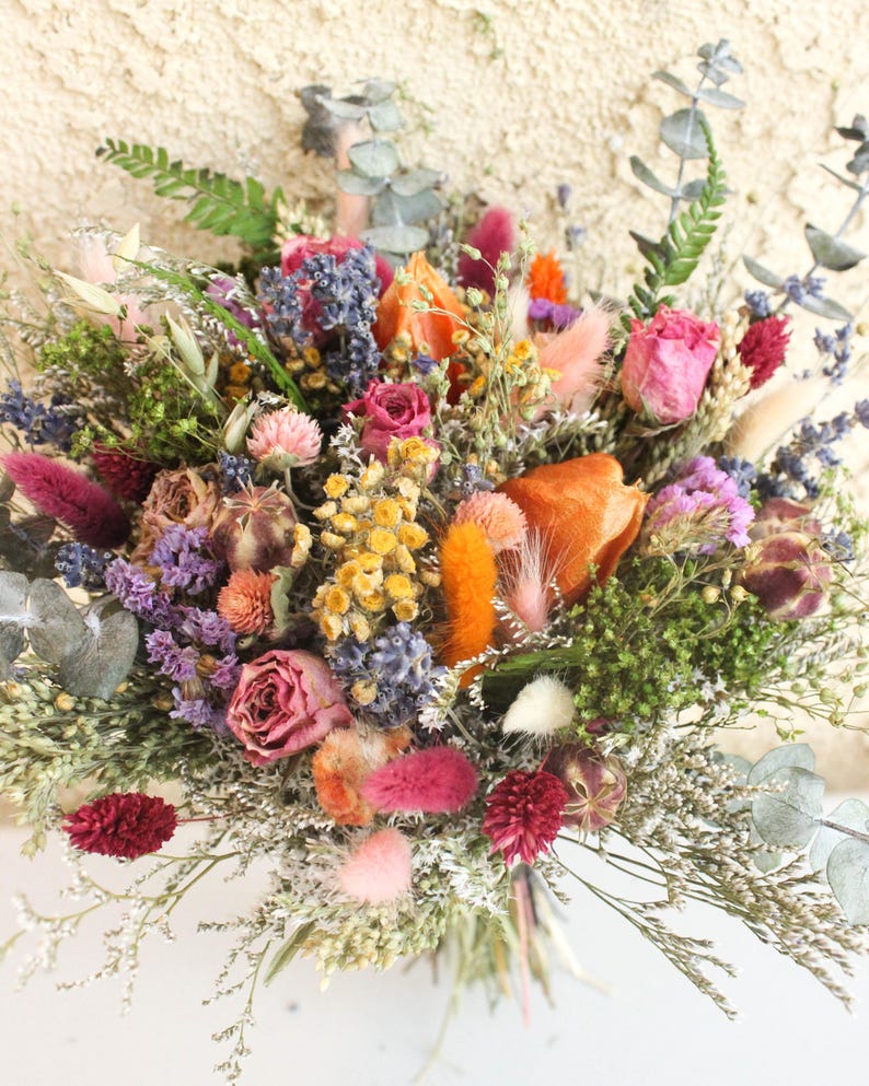 May include: A vibrant bouquet of dried flowers featuring a mix of colors including pink, orange, yellow, and purple. The arrangement includes roses, lavender, and various types of foliage. The bouquet is tied together, creating a natural and rustic aesthetic.