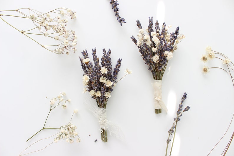 May include: Three small bouquets of dried lavender and white flowers tied with twine. The bouquets are arranged on a white surface.