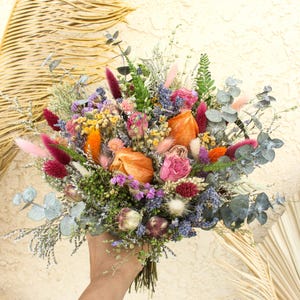 May include: A vibrant bouquet of dried flowers featuring a mix of colors including orange, pink, purple, and blue. The bouquet includes various textures and shapes, with eucalyptus leaves and feathery accents. The flowers are held together with a natural twine.