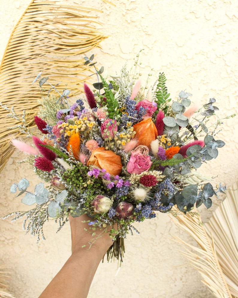 May include: A vibrant bouquet of dried flowers featuring a mix of colors including orange, pink, purple, and blue. The bouquet includes various textures and shapes, with eucalyptus leaves and feathery accents. The flowers are held together with a natural twine.