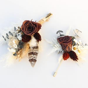 May include: Two boutonnieres with dried flowers and feathers. One boutonniere features a brown rose, white flowers, and a feather with brown and white stripes. The other boutonniere has a brown rose, red accents, and a gold pin.