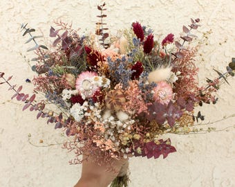 Puede incluir: Un ramo de flores secas con una mezcla de tallos en tonos burdeos, rosa y azul. El arreglo incluye eucalipto, lavanda, colas de conejo, siemprevivas y una cápsula de amapola central, todo atado en la base con cuerda de fibra natural.
