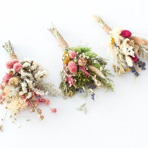 May include: Three dried flower bouquets with a variety of textures and colors. Each bouquet is tied with twine. The flowers include pink, white, yellow, and purple blooms, along with dried grasses and greenery, set against a white background.