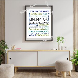 May include: A framed art print with a white background and black frame. The print features words like "Unstoppable," "Jeremiah," and "Champion" in blue and green fonts. The artwork is displayed on a white cabinet with wooden legs, with plants and decor.
