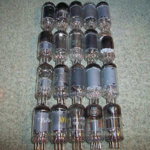 Pode incluir: Uma coleção de válvulas de vácuo vintage, dispostas em fileiras. As válvulas são feitas de vidro transparente com vários componentes internos visíveis. Algumas válvulas têm tampas cinzas ou pretas, e algumas têm texto impresso. O fundo é um tapete verde.