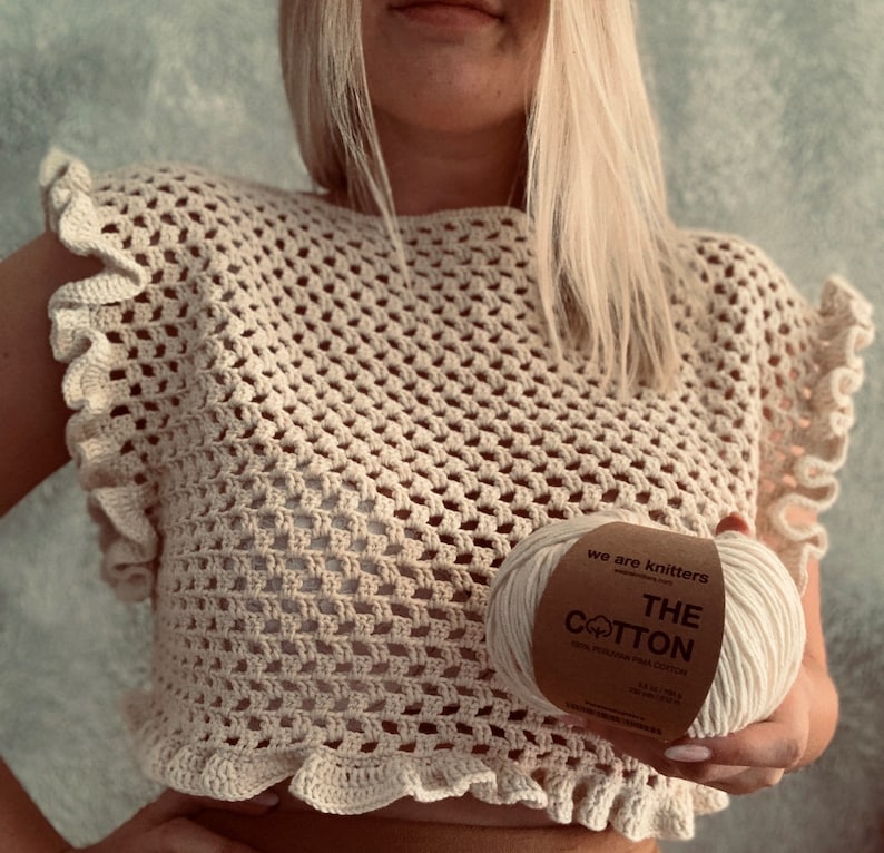 May include: A close-up of a crocheted top with a ruffled neckline. The top is made with a beige yarn and has a loose, open weave. A ball of yarn is in the foreground, with the label "We are knitters The Cotton 100% Peruvian Pima Cotton" visible.