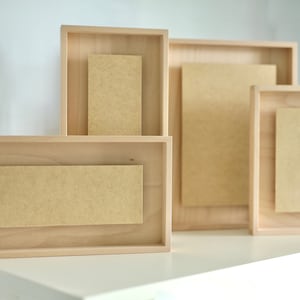 Könnte beinhalten: Vier Holzbilderrahmen in verschiedenen Größen, alle mit einer hellbraunen Oberfläche. Die Rahmen sind in einer Reihe angeordnet, wobei der größte Rahmen hinten und der kleinste Rahmen vorne ist.