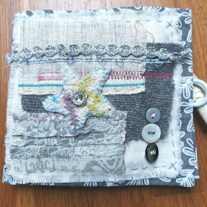 Puede incluir: Un diario de tela hecho a mano con un diseño de patchwork. La cubierta presenta un adorno en forma de estrella con un centro de pedrería, telas estampadas grises y blancas y costuras decorativas. Tres botones están cosidos en la cubierta.