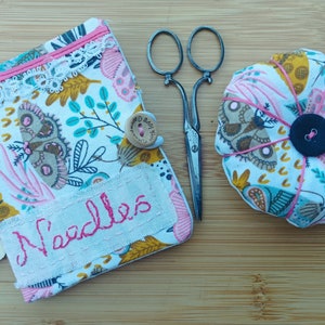 May include: A white fabric needle case with a pink zipper, lace trim, and embroidered butterflies. The case has a pink fabric flap with the word "Needles" embroidered in pink thread. The case is next to a pink spool of thread, a pincushion with a pink button and butterflies, and a pair of antique scissors.