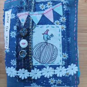May include: A handcrafted denim book with a patchwork design. The book features a central square with a drawing of a bird on a ball of yarn, surrounded by floral and banner accents. The text "LOVE TO KNIT" is visible on a white label.