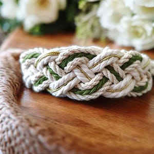 May include: Braided bracelet featuring a cream-colored rope design with green and gold accents. The bracelet is made of woven rope and has a nautical aesthetic. The bracelet is displayed on a wooden surface with a bouquet of white flowers in the background.