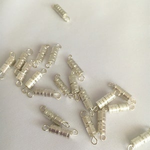 Pode incluir: Fechos de metal prateados para fazer joias. Os fechos são cilíndricos com uma superfície texturizada e têm um pequeno laço em cada extremidade.