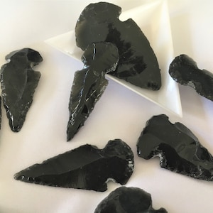 May include: A collection of eight black obsidian arrowheads, some with a slight green tint, arranged on a white surface.