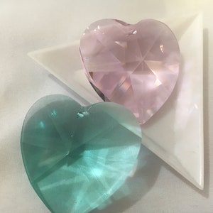May include: Two heart-shaped crystal prisms, one pink and one teal green, on a white triangular surface.