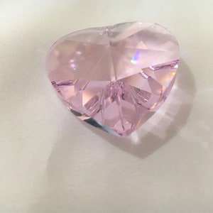 May include: A pink crystal heart shaped prism with many facets. The prism is sitting on a white surface.