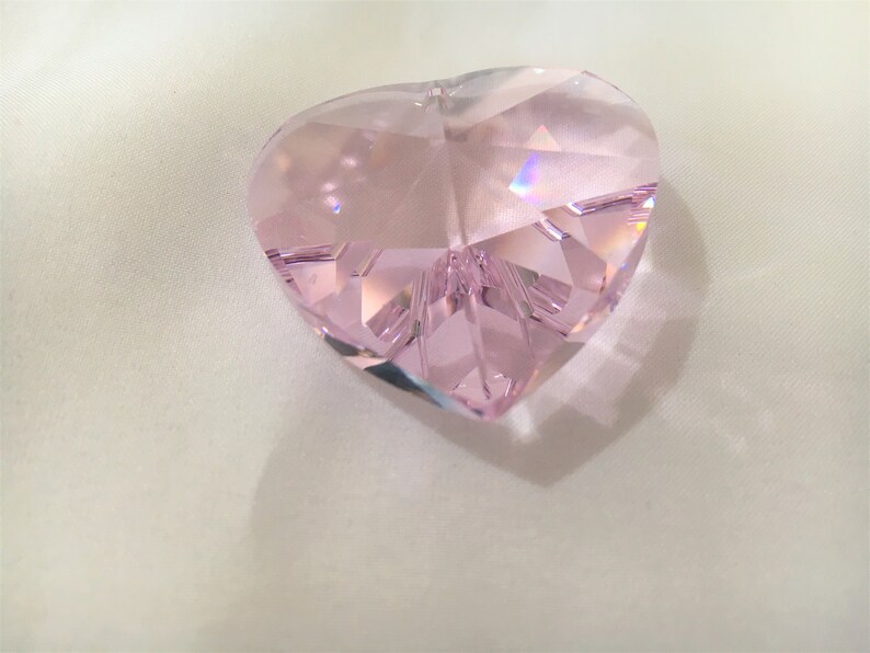 May include: A pink crystal heart shaped prism with many facets. The prism is sitting on a white surface.
