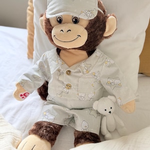 Op de afbeelding: Een pluche aap in een lichtgrijze pyjamaset met een patroon van gele sterren en witte eendjes. De aap draagt een bijpassend slaapmasker over de ogen en houdt een kleine witte pluche beer vast. De pyjama is voorzien van twee bruine knopen.