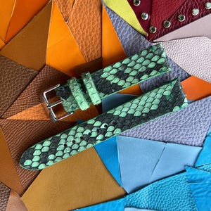 Könnte beinhalten: Zweiteiliges Uhrenarmband mit grünem und schwarzem Schlangenmuster, mit silberner Schnalle. Das Armband wird auf einem farbenfrohen Hintergrund aus verschiedenen Lederstücken in Orange-, Braun-, Blau- und Lilatönen präsentiert.