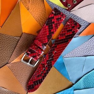May include: Red and black snakeskin-patterned watch straps with a silver buckle, displayed on a colorful patchwork of leather pieces in shades of orange, brown, blue, and red. The straps are new and unused.