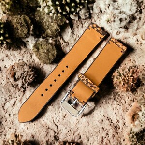 May include: Two brown leather watch straps with a textured pattern. The straps have stitching around the edges and are laid out on a textured surface.