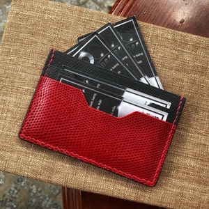 May include: A red and black leather card holder with a textured finish. The card holder is filled with several black and white business cards. The card holder is resting on a textured, tan surface.