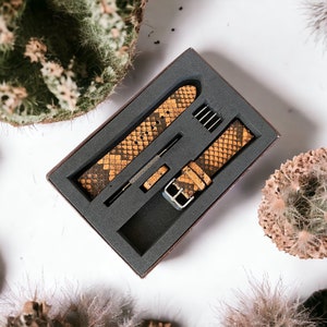 May include: Two brown and yellow patterned watch bands in a black box with a gray foam interior. The bands are made of leather and have a snakeskin pattern.