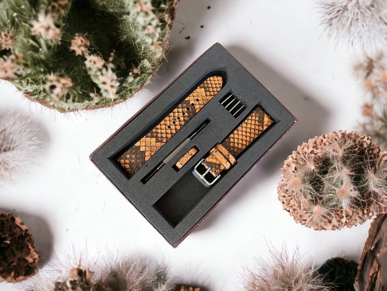 May include: Two brown and yellow patterned watch bands in a black box with a gray foam interior. The bands are made of leather and have a snakeskin pattern.