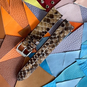 Könnte beinhalten: Ein Uhrenarmband mit Schlangenmuster in Braun- und Beigetönen, mit einer silbernen Schnalle. Es liegt auf einem bunten Hintergrund aus verschiedenen Lederstücken in Orange, Blau, Rosa und Rot.