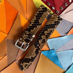 Peut inclure: Un bracelet de montre à motif peau de serpent marron et noir, avec une boucle argentée. Le bracelet est présenté sur un fond coloré d'échantillons de cuir dans diverses nuances d'orange, de rouge et de bleu.