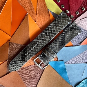 Op de afbeelding: Een horlogebandje met een slangenpatroon in grijstinten en zwart, met een zilveren gesp. De band wordt getoond op een kleurrijke achtergrond van leren stukken in verschillende kleuren, waaronder oranje, rood en blauw.