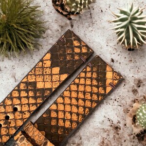 May include: Two brown leather watch straps with a snakeskin pattern and gold accents. The straps are laid out on a light gray surface.