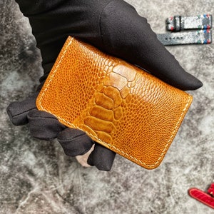May include: A brown leather card holder with a textured ostrich skin pattern. The card holder has white stitching around the edges.