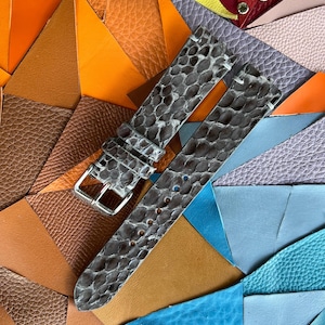 Può includere: Cinturini grigi per orologi con motivo a pelle di rettile e fibbia argentata. I cinturini sono esposti su uno sfondo colorato di pezzi di pelle in vari colori e trame, tra cui arancione, marrone e blu.