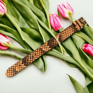May include: A brown leather watch band with a snakeskin pattern. The band is lying on a white surface with pink tulips.