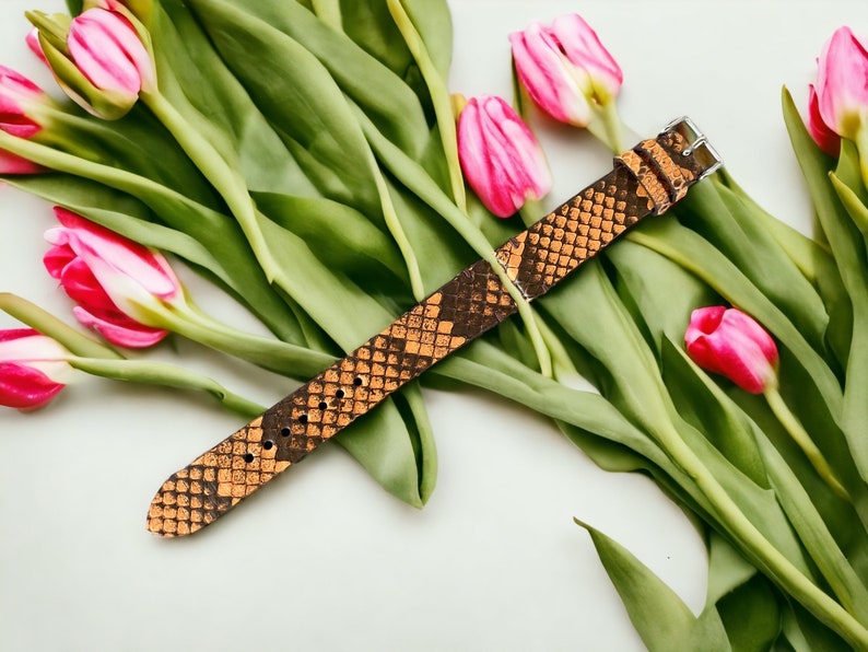 May include: A brown leather watch band with a snakeskin pattern. The band is lying on a white surface with pink tulips.