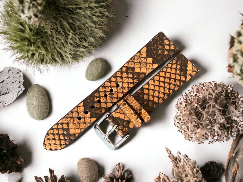 May include: A brown leather watch strap with a snakeskin pattern. The strap is laid out on a white surface with small decorative rocks and plants.