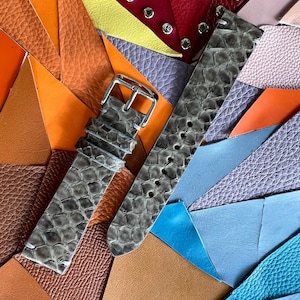 Puede incluir: Una correa de reloj gris con estampado de piel de serpiente y hebilla plateada descansa sobre un colorido mosaico de muestras de cuero. Las piezas de cuero son en tonos naranja, marrón, azul, morado y rojo, creando un fondo vibrante.