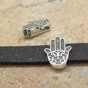 Può includere: Due ciondoli a forma di mano di hamsa argentati con disegni intricati. I ciondoli misurano circa 2,5 cm e presentano un motivo dettagliato e ornato.
