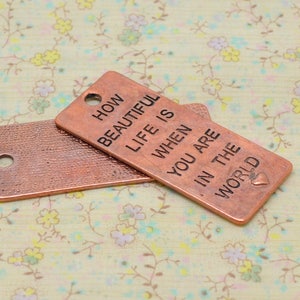 Peut inclure: Deux petites étiquettes rectangulaires en cuivre avec une surface texturée. L'étiquette du haut porte le texte "How beautiful life is when you are in the world" gravé dessus. L'étiquette du bas est vide.