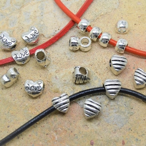 May include: A collection of silver-toned beads with various designs, including hearts with "love" inscribed, striped shields, and heart-shaped beads. The beads are strung on red and black leather cords, set against a neutral background.