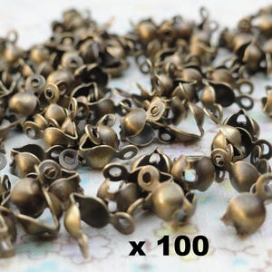 Peut inclure: Une grande quantité de petites cloches en métal doré antique avec une boucle sur le dessus. Les cloches sont disposées de manière aléatoire sur une surface blanche. Le texte "x 100" est imprimé en noir sur la surface.