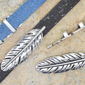 May include: Two silver feather-shaped belt buckles, one on a blue denim strap and the other on a black leather strap. A third silver buckle with a straight edge is also shown.