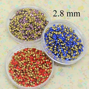 May include: Three containers of small, faceted rhinestones. The containers are clear plastic and the rhinestones are red, blue, and purple. The text "2.8 mm" is printed on the image.